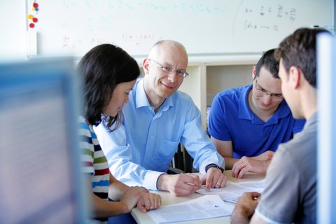 Bild von einem Dozenten und 3 Studierenden, die um einen Tisch sitzen und gemeinsam ein Paper durchsprechen. Im Hintergrund sind ein Holzregal und ein Whiteboard zu erkennen.