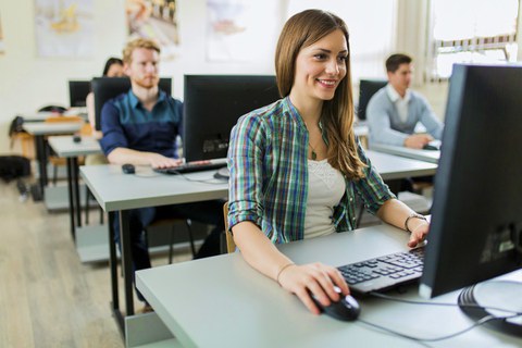 Das Foto zeigt mehrere Personen, die in einem Computerraum sitzen und an ihren Computern arbeiten. Im Vordergrund sitzt eine junge, lächelnde Frau.