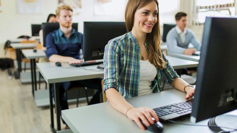 Das Foto zeigt mehrere Personen, die in einem Computerraum sitzen und an ihren Computern arbeiten. Im Vordergrund sitzt eine junge, lächelnde Frau.