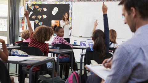 Das Foto zeigt eine Referendarin beim Unterrichten einer Grundschulklasse. Viele Lernende melden sich. Im Vordergrund sitzt der Lehrer und macht sich Notizen auf einem Klemmbrett. 