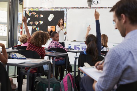 Das Foto zeigt eine Referendarin beim Unterrichten einer Grundschulklasse. Viele Lernende melden sich. Im Vordergrund sitzt der Lehrer und macht sich Notizen auf einem Klemmbrett. 