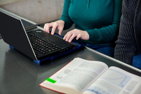 Foto: Eine Frau schreibt an einem Laptop mit Braille-Zeile. Ein Buch liegt auf dem Tisch.