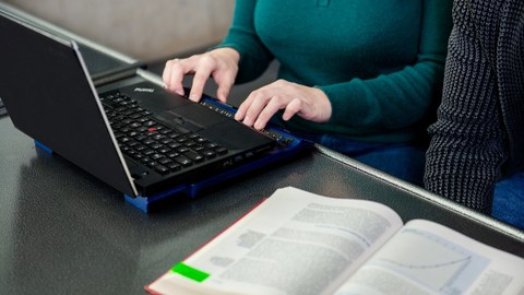Foto: Eine Frau schreibt an einem Laptop mit Braille-Zeile. Ein Buch liegt auf dem Tisch.