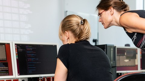 Foto: Eine Studentin und eine Dozentin beugen sich über einen Monitor in einem Computerkabinett.