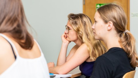 Foto von drei Studentinnen, die in einem Seminar an einem Tisch sitzen und zuhören.
