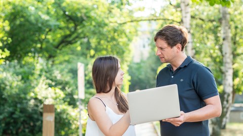 Foto: Eine Frau und ein Mann halten einen Laptop in der Hand und unterhalten sich im Freien.
