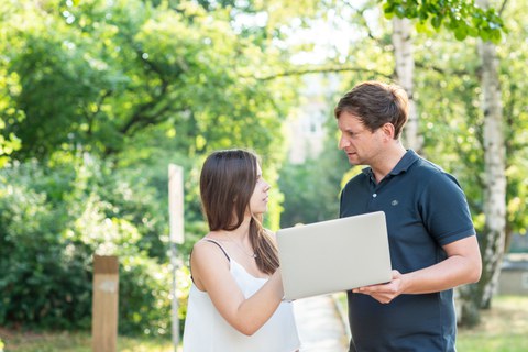 Foto: Eine Frau und ein Mann halten einen Laptop in der Hand und unterhalten sich im Freien.