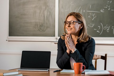 Foto: Das Foto zeigt eine Lehrerin an ihrem Schreibtisch in einem Klassenzimmer.