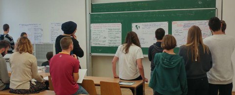 Bild mehrerer Studierender, die vor einer Tafel und einer Wand stehen und die Plakate betrachten, die daran angebracht sind. 