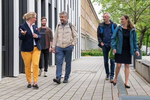 Foto: 4 Personen gehen nebeneinander auf einem Gehweg neben einem Gebäude auf den:die Betrachter:in zu.