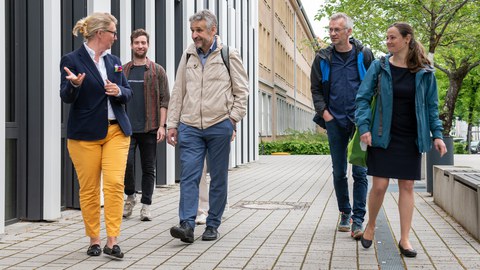 Foto: 4 Personen gehen nebeneinander auf einem Gehweg neben einem Gebäude auf den:die Betrachter:in zu.