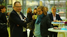 Bild, auf dem ein Mann und eine Frau an einem Stehtisch stehen. Letztere trinkt aus einer Tasse. Im Hintergrund sind weitere Tische und Personen  zu sehen.