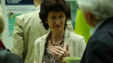 Bild einer Frau, die sich im Gespräch mit einem Mann befindet. Im Hintergrund sind Informations-Grafiken zu sehen, vor denen weitere Personen stehen.