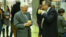 Bild, auf dem zwei Männer einander gegenüber stehen. Beide halten eine Tasse in der Hand. Der linke Mann lacht, der rechte setzt gerade zum trinken an. Im Hintergrund sind weitere Personen und Informationstafeln zu sehen.