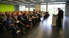Bild von einem Hörsaal mit Stuhlreihen, der voll besetzt zu sein scheint. Das Publikum hört einem Mann zu, der vorne an einem Pult mit Mikrofon steht und augenscheinlich einen Vortrag hält.