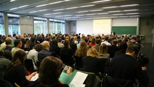 Bild von einem Hörsaal mit Stuhlreihen, der voll besetzt zu sein scheint. Das Publikum hört einer Person zu, die vorne vor einer Tafel an einem Pult steht und augenscheinlich einen Vortrag hält. Links von der Tafel wird etwas an die Wand projiziert.