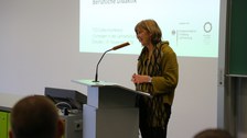 Bild von einer Frau, die vor einer Tafel an einem Pult mit Mikrofon steht und spricht. Links von der Tafel wird eine Präsentation an die Wand projiziert.