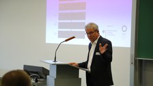Bild von einem Mann, der vor einer Tafel an einem Pult mit Mikrofon steht und spricht. Links von der Tafel wird eine Präsentation an die Wand projiziert.