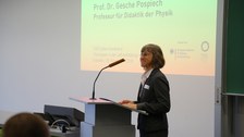 Bild von einer Frau, die vor einer Tafel an einem Pult mit Mikrofon steht und spricht. Links von der Tafel wird eine Präsentation an die Wand projiziert.