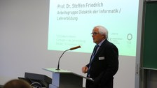 Bild von einem Mann, der vor einer Tafel an einem Pult mit Mikrofon steht und spricht. Links von der Tafel wird eine Präsentation an die Wand projiziert.