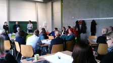 Bild eines Seminarraumes im Hörsaalzentrum der TU Dresden, in dem im Vordergrund mehrere Personen an verschiedenen Gruppentischen sitzen. Im Hintergrund stehen 3 Menschen vor einer Tafel und präsentieren.