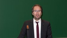 Bild von einem Mann, der vor einer Tafel an einem Pult mit Mikrofon steht und spricht.