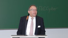 Bild von Prof. Axel Gehrmann. Er steht an einem Pult mit Mikrofon vor einer Tafel und spricht. An der Tafel steht "#TUDSylber".