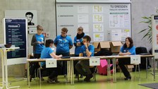 Links im Bild steht ein Flipchart mit einem Tagesprogramm. Daneben sind drei Tische aufgereiht, an denen jeweils ein Schild hängt, mit jeweils einer Person mit blauem T-Shirt dahinter. Drei weitere gleich gekleidete Personen stehen dahinter.