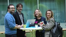 Bild von zwei Männern und zwei Frauen, die um einen Stehtisch herum stehen und in die Kamera lächeln. Auf dem Tisch steht ein Laptop, der von einem der Männer bedient wird.