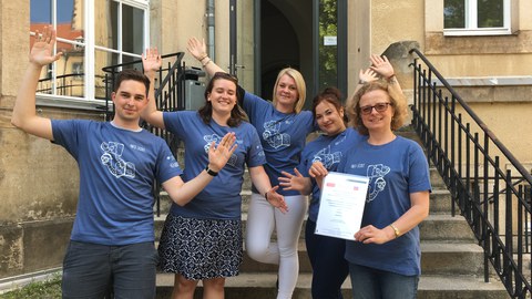 Bild einer Gruppe von 5 Personen, die auf einer Treppe stehen und in die Kamera lächeln. Sie tragen alle das gleiche blaue T-Shirt mit der weißen Aufschrift "Info-Scout". Die rechts stehende Frau präsentiert eine Urkunde.