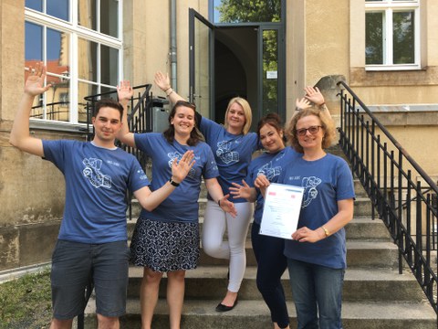 Bild einer Gruppe von 5 Personen, die auf einer Treppe stehen und in die Kamera lächeln. Sie tragen alle das gleiche blaue T-Shirt mit der weißen Aufschrift "Info-Scout". Die rechts stehende Frau präsentiert eine Urkunde.