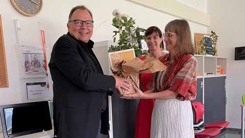 Foto: drei Personen stehen in einem Raum. Einer Person wird ein Geschenk übergeben.