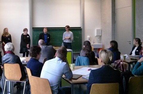 Bild eines Seminarraumes am Hörsaalzentrum der TU Dresden. Im Vordergrund sitzen einige Erwachsene an verschiedenen Gruppentischen. Sie hören den vier Präsentierenden zu, welche nebeneinander vor der Tafel stehen und reden.