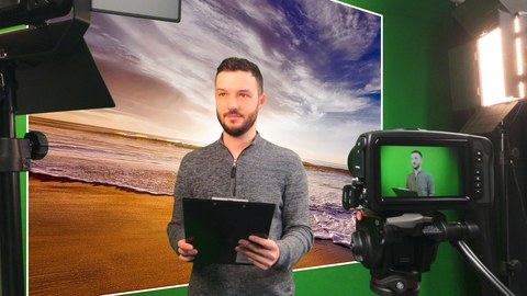 Foto: 2 Scheinwerfer und eine Kamera sind auf eine Person vor einem Greenscreen gerichtet, wobei als Hintergrund ein digitales Bild von einem Sonnenuntergang am Meer eingefügt ist.