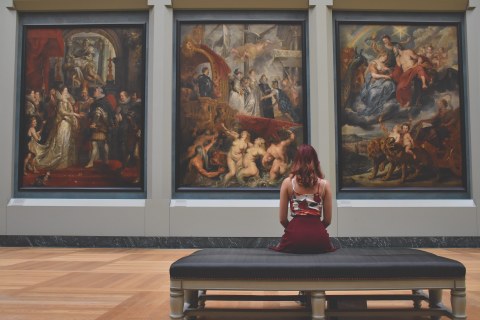 Foto einer Frau, die in einem Museum auf einem Sofa sitzt und ein Triptychon betrachtet.