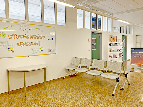 Foto eines Ganges im Seminargebäude 2. Im Vordergrund sind Sitzbänke und eine Tafel mit der Aufschrift "Studienbüro Lehramt" zu sehen. Im Hintergrund sind ein Aufsteller, Flyer und zwei Türen zu erkennen.