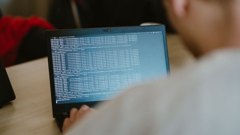 Foto zeigt Jugendlichen von hinten, während er an einem Laptop sitzt, welcher Code anzeigt.