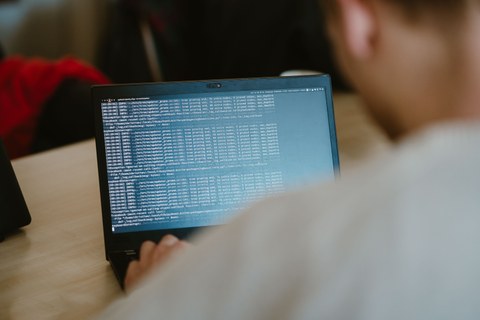 Foto zeigt Jugendlichen von hinten, während er an einem Laptop sitzt, welcher Code anzeigt.