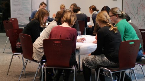Bild von einem Raum, in dem mehrere Personen um zwei Tische herum verteilt sitzen und miteinander sprechen. Im Hintergrund sind mehrere beschriebene Flipchart-Blätter zu sehen.