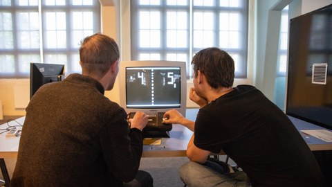 Zwei Männer sitzen vor einem Bildschirm und spielen gemeinsam das Videospiel "Pong" 