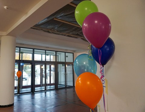 Auf dem Foto sind fünf verschiedenfarbige Luftballons zu sehen, die an einer weißen Wand im Vordergrund hängen. Im Hintergrund ist eine Glaseingangstür und das Foyer zu sehen.