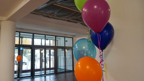 Auf dem Foto sind fünf verschiedenfarbige Luftballons zu sehen, die an einer weißen Wand im Vordergrund hängen. Im Hintergrund ist eine Glaseingangstür und das Foyer zu sehen.