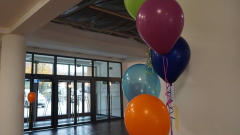 Auf dem Foto sind fünf verschiedenfarbige Luftballons zu sehen, die an einer weißen Wand im Vordergrund hängen. Im Hintergrund ist eine Glaseingangstür und das Foyer zu sehen.