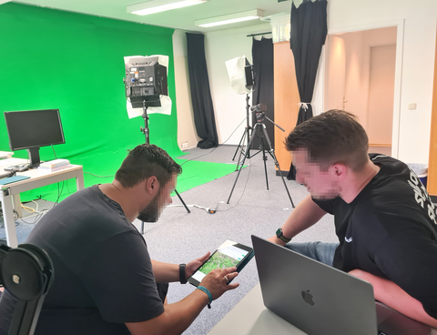 Zwei Männer arbeiten zusammen an einem Tablet. Im Hintergrund ist ein Medienlabor mit Green Screen zu sehen.