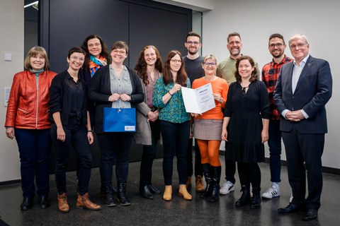 Gruppenfoto mit 12 Personen im Rahmen einer Preisverleihung. Zwei Frauen im Vordergrund halten eine Urkunde.