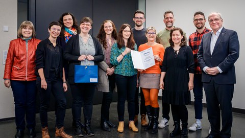 Gruppenfoto mit 12 Personen im Rahmen einer Preisverleihung. Zwei Frauen im Vordergrund halten eine Urkunde.