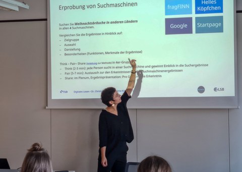 Frau hält Vortrag im Seminarraum und zeigt auf eine Leinwand, auf der eine Präsentation gezeigt wird.