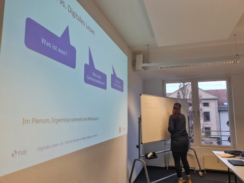 Frau schreibt was auf ein Whiteboard, zu sehen ist außerdem eine Präsentation auf einer Leinwand.