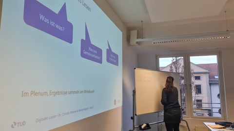 Frau schreibt was auf ein Whiteboard, zu sehen ist außerdem eine Präsentation auf einer Leinwand.