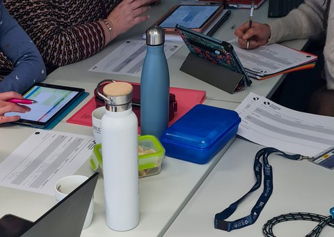 Tisch mit Arbeitsmaterialien wie Tablets, Stiften und Getränkeflaschen.